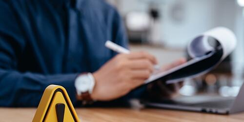 exclamation point on desk, with businessman in background making notes on a clipboard